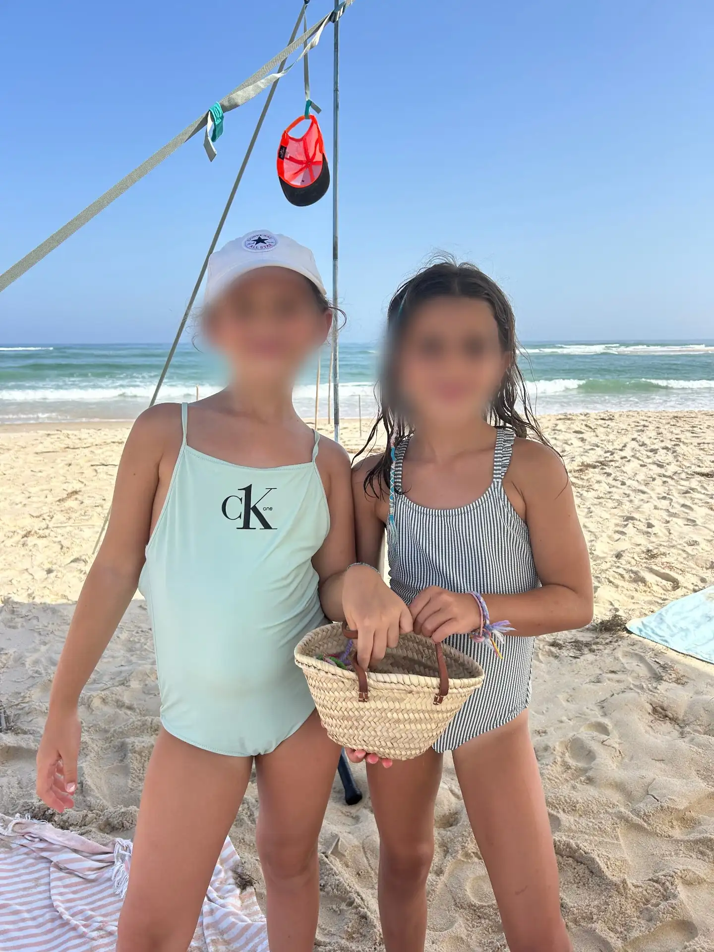 Mimi and Aurora standing on the sand, holding a woven palm basket, with the sea behind them