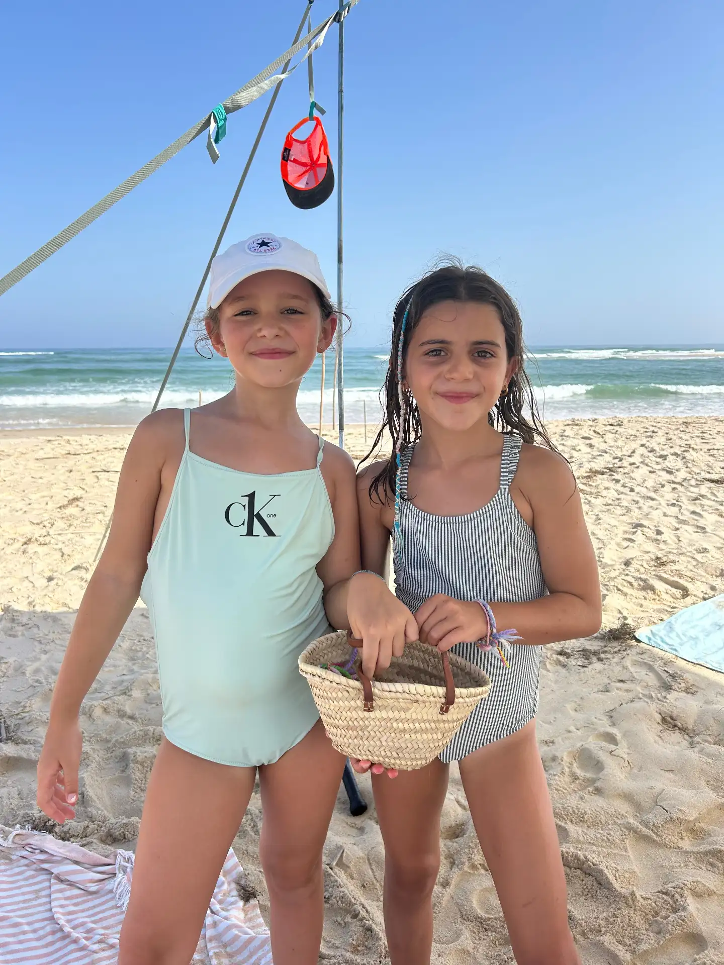 Mimi and Aurora standing on the sand, holding a woven palm basket, with the sea behind them