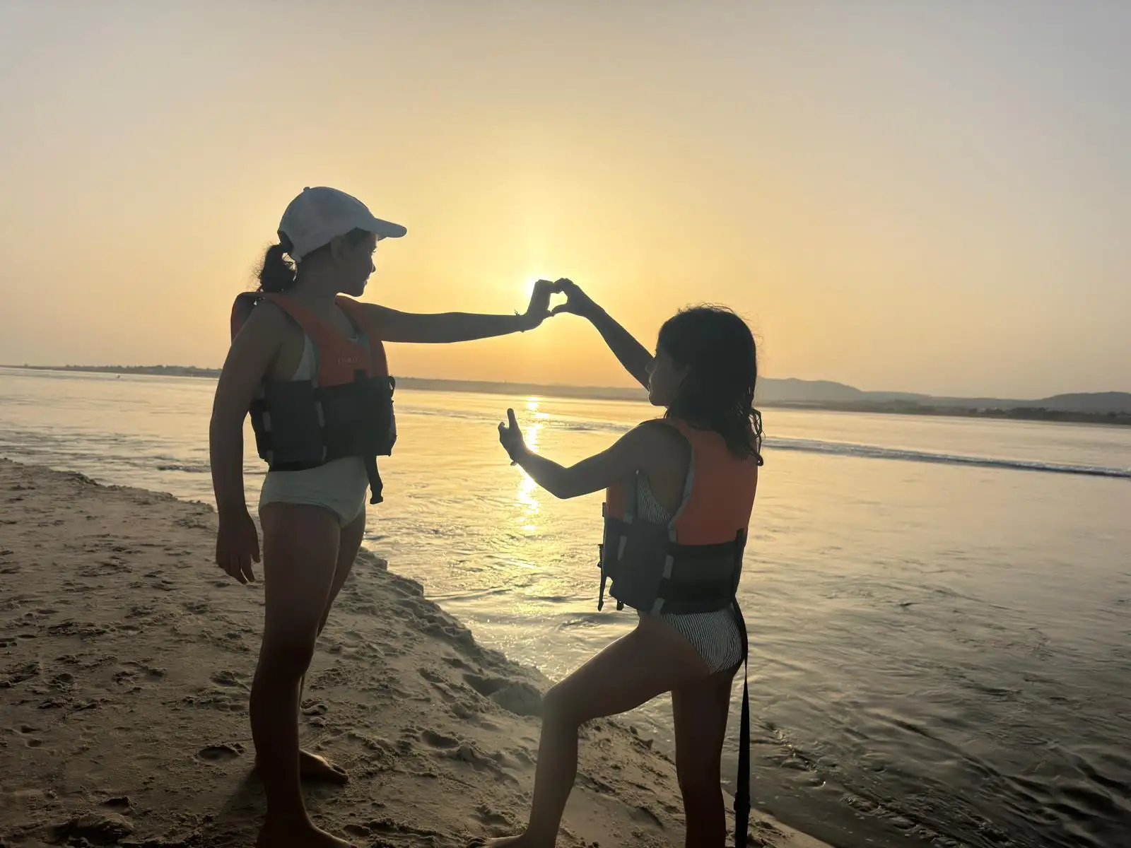 Two friends holding hands in a heart shape at sunset